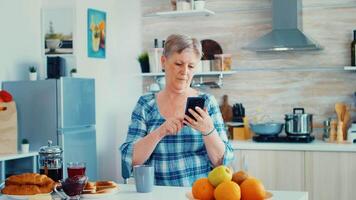 Single senior woman surfing on phone in kitchen during breakfast. Authentic elderly person using modern smartphone internet technology. Online communication connected to the world, senior leisure time with gadget at retirement age video