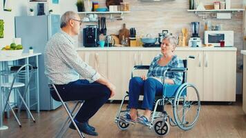 Disabled woman sitting in wheelchair having a conversation with elderly husband in kitchen. Old man having a talking with wife. Living with handicapped person with walking disabilities video