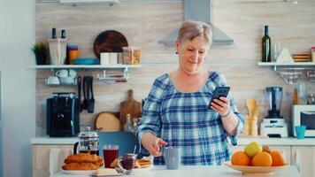 Senior mulher desfrutando música a partir de Smartphone durante café da manhã dentro cozinha segurando copo do café. relaxado idosos dançando, Diversão estilo de vida com moderno tecnologia video