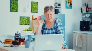 Oma winken während ein Video Konferenz mit Laptop während haben Frühstück im Küche. Alten Person mit Internet online Plaudern Technologie Video Webcam Herstellung ein Video Anruf Verbindung Kamera Kommunikation Konferenz Anruf