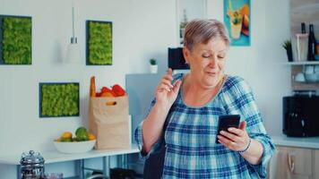 contento anziano donna ascoltando musica nel cucina su smartphone durante colazione sabato. rilassato anziano ballare, divertimento stile di vita con moderno tecnologia video