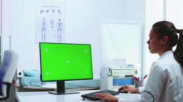 Doctor using computer with green screen display in hospital cabinet and assistant in the background. Medic in white coat working on monitor with chroma key in clinic cabinet to check patient diagnose. video