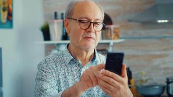 Old man surfing on social media using smartphone during breakfast in kitchen. Authentic portrait of retired senior enjoying modern internet online technology video