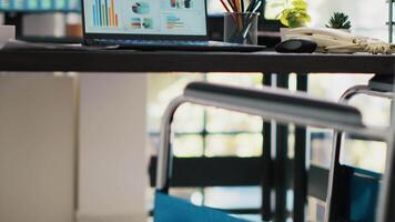 Wheelchair in inclusive office for employees and laptop on desk showing statistics. Medical chair in workspace offering accessibility to workers with disability and notebook with financial charts video