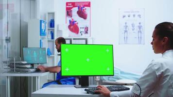 asistente dando médico radiografía imagen mientras trabajando en computadora con verde pantalla monitor en hospital gabinete. escritorio con reemplazable pantalla en médico clínica mientras médico es comprobación paciente radiografía para diagnosticar. video