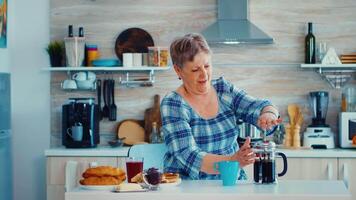 Mature woman using french press to make coffee in kitchen during breakfast. Elderly person in the morning enjoying fresh brown cafe espresso cup caffeine from vintage mug, filter relax refreshment video