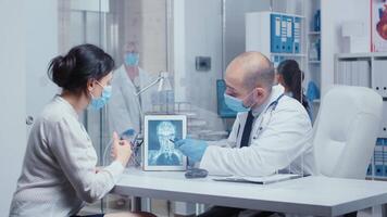 Healthcare diagnosis during COVID-19 for woman patient who looks at X Ray on a digital tablet through a plexiglass wall. Medical consultation in protective equipment concept shot of sars-cov-2 global health pandemic crisis video