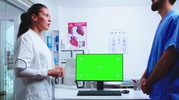 Doctor analysing patient results with assistant on computer with green screen in hospital. Medic wearing white coat in clinic pointing at desktop with chroma key talking with nurse about patient diagnose video