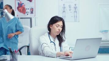 Young woman doctor in private medical cabinet typing on laptop while the nurse in working in the background. Healthcare system medical worker practitioner in hospital and health research video