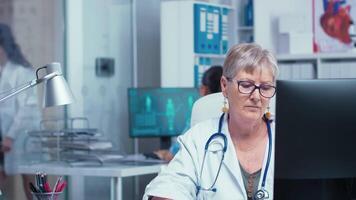 retrato do Senior médico mulher trabalhando em pc dentro moderno privado gabinete enquanto a enfermeira é verificação x raios dentro a costas e de outros médico coisa anda em dentro corredor. consultando dentro cuidados de saúde sistema video