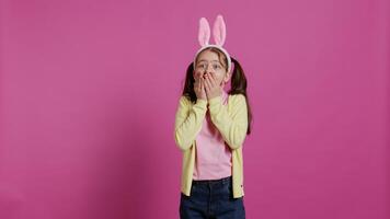 adorable surpris écolière étant dans choc après voyant quelque chose dans studio, souriant et sentiment très heureux. joyeux petit enfant avec lapin oreilles étant étonné par cadeaux. caméra b. video