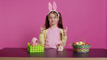 pequeño alegre niño posando con dedos cruzado en estudio, sentado a un mesa lleno con Pascua de Resurrección festivo adornos joven Chica de escuela esperando para suerte y fortuna durante primavera fiesta celebracion. cámara b. video