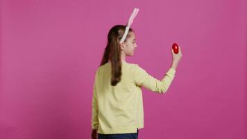 Enthusiastic small child spinning around in front of camera, presenting her red painted egg for easter sunday festivity. Cheery toddler with bunny ears twirling and doing a pirouette. Camera B. video