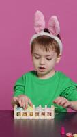 Vertical Video Happy pleased child decorating easter arrangements with handcrafted items, arranging painted colorful eggs in a basket. Joyful little boy celebrating spring festivity, pink backdrop. Camera A.