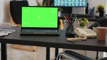 Focus on green screen laptop on office desk with computer in blurry background showing stock exchange data. Close up of mockup notebook and trading platform displayed on PC screen in the back video