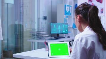 médico leyendo en tableta ordenador personal con verde pantalla en hospital gabinete acerca de un peligroso virus. asistente en azul médico uniforme. médico en salud clínica trabajando en tableta computadora con reemplazable pantalla haciendo medicina investigación. video