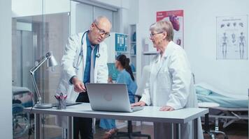 Two elderly senior experienced doctors deciding patient treatment while nurse is working in background. Senior authentic doctors in modern private hospital clinic room healthcare system medicine and treatment video