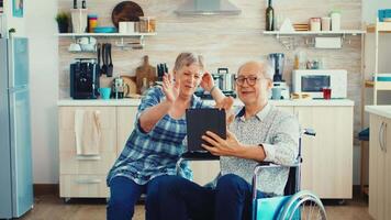 senior paar lachend en golvend gedurende een video telefoontje met kleinkinderen gebruik makend van tablet computer in keuken. verlamd gehandicapten oud ouderen Mens gebruik makend van modern communicatie technologie.