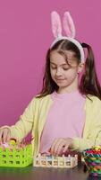 Vertical Video Smiling happy schoolgirl presenting a handmade decorated basket in studio, making easter holiday preparations against pink background. Young child showing handcrafted arrangements. Camera A.