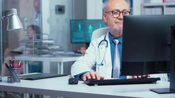 Elderly doctor working on PC in modern private clinic with glass walls. Hospital professionals in health industry staff at work in medicine medical practitioner specialist in unifor at desk job video