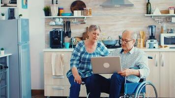 Desativado Senior homem dentro cadeira de rodas e esposa navegando em Internet usando computador portátil dentro cozinha. paralisado deficiente velho idosos homem usando moderno comunicação tecnologia. video