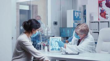 Senior woman doctor showing a X ray on tablet in times of COVID-19, wearing mask and protection gloves and talking through a plexiglass wall. Medical consultation in protective equipment concept shot of sars-cov-2 global health pandemic crisis video