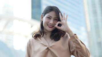 Teenager Asian woman feeling happy smiling and looking to camera while relax at outdoor city space. Portrait of successful business woman showing OK hand and wave up hand. Happy feeling concept. video