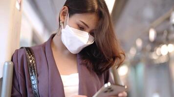 asiatisk företag kvinna bär ansikte mask medan reser i massa genomresa tåg. kvinna är tittar på något på smart telefon med medan stående på fönster. skydd för epidemi och hälsa i offentlig. video