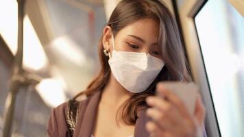 asiatico attività commerciale donna indossare viso maschera mentre in viaggio nel massa transito treno. femmina è Guardando qualcosa su inteligente Telefono con mentre in piedi a finestra. protezione per epidemico e Salute nel pubblico. video