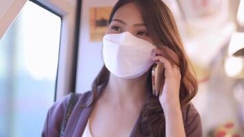 asiatico attività commerciale donna indossare viso maschera mentre in viaggio nel massa transito treno. femmina è Guardando qualcosa su inteligente Telefono con mentre in piedi a finestra. protezione per epidemico e Salute nel pubblico. video