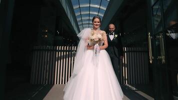 joyeux la mariée et jeune marié sortir le bâtiment, une radieux la mariée avec une bouquet des promenades en dehors le porte, suivi par une souriant jeune marié sur une ensoleillé journée. video