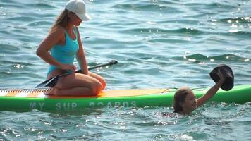 mar mujer sorber. silueta de contento positivo joven mujer, madre y hija en azul bikini, surf en verde cenar tablero mediante calma agua superficie en puesta de sol. activo estilo de vida a mar río. lento movimiento video