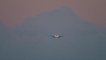 lange Schuss von ein Jet Flugzeug mit ein unkenntlich Lackierung mit Scheinwerfer auf Annäherung zum Landung im das Sonnenuntergang wolkig Himmel video