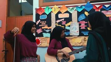 Three women in a classroom setting, one playing guitar, with colorful paper decorations on the wall. video