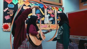 Three women in a classroom setting, one playing guitar, with colorful paper decorations on the wall. video