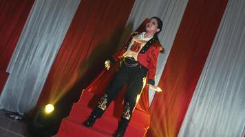 Person in a vampire costume posing dramatically on red stairs with a cape and fangs, against a draped curtain background. video