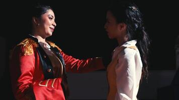 Two women in theatrical costumes on stage, one in a red jacket, engaging in a dramatic scene. video