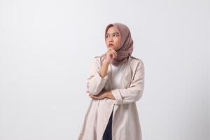 Portrait of confused Asian hijab woman in casual suit thinking with hand on chin, feeling doubt while making choice. Businesswoman concept. Isolated image on white background photo