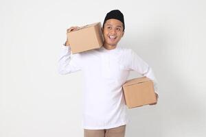 Portrait of attractive Asian muslim man in koko shirt with peci feeling grateful and peacful while carrying cardboard box. Going home for Eid Mubarak. Isolated image on white background photo