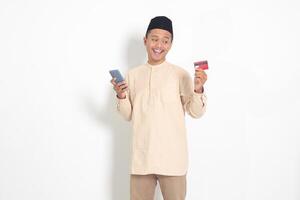 Portrait of attractive Asian muslim man in koko shirt with skullcap holding a mobile phone and presenting credit card. Isolated image on white background photo