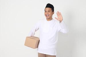 Portrait of excited Asian muslim man in koko shirt with peci carrying cardboard box while saying hi, greeting to someone. Going home for Eid Mubarak. Isolated image on white background photo