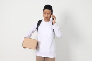 Portrait of worried Asian muslim carrying cardboard box while having having call on mobile phone, showing sad expression. Going home for Eid Mubarak. Isolated image on white background photo