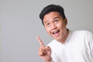 Portrait of Asian muslim man in white koko shirt poiting to the side and showing wow face expression. Isolated image on gray background photo