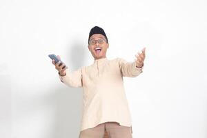 Portrait of surprised Asian muslim man in koko shirt with skullcap holding mobile phone, showing wow shocked face expression. Advertising and social media concept. Isolated image on white background photo