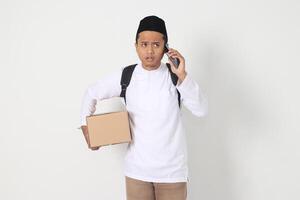 Portrait of worried Asian muslim carrying cardboard box while having having call on mobile phone, showing sad expression. Going home for Eid Mubarak. Isolated image on white background photo