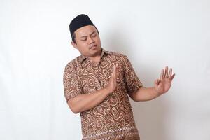 retrato de desagradable asiático hombre vistiendo batik camisa y Songkok formando un mano gesto a evitar algo. publicidad concepto. aislado imagen en gris antecedentes foto
