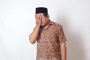 Portrait of suffering Asian man wearing batik shirt and songkok having headache while touching his forehead area. Isolated image on gray background photo