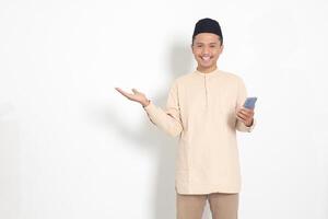 Portrait of attractive Asian muslim man in koko shirt with skullcap holding mobile phone, pointing and showing product with finger. Social media concept. Isolated image on white background photo