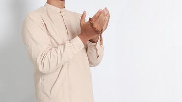 Close up portrait of religious Asian muslim man in koko shirt with skullcap praying earnestly with his hands raised, holding islamic beads. Devout faith concept. Isolated image on white background photo