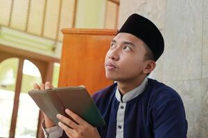 Religious Asian man in muslim shirt and black cap reading the holy book of Quran in the public mosque photo